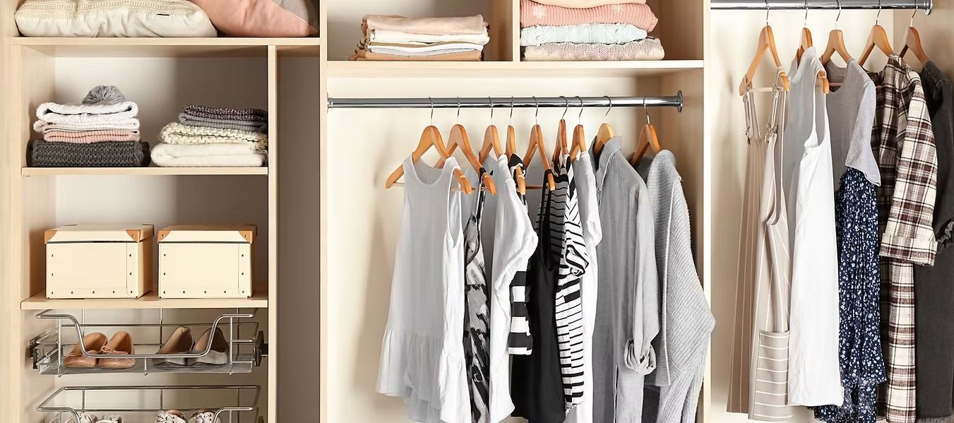 A well-organized closet with clothing on hangers and folded on shelves. The items are in neutral colors, creating a minimalist feel.