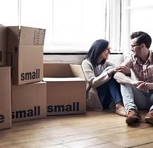 A couple sits on a wooden floor, surrounded by cardboard boxes labeled
