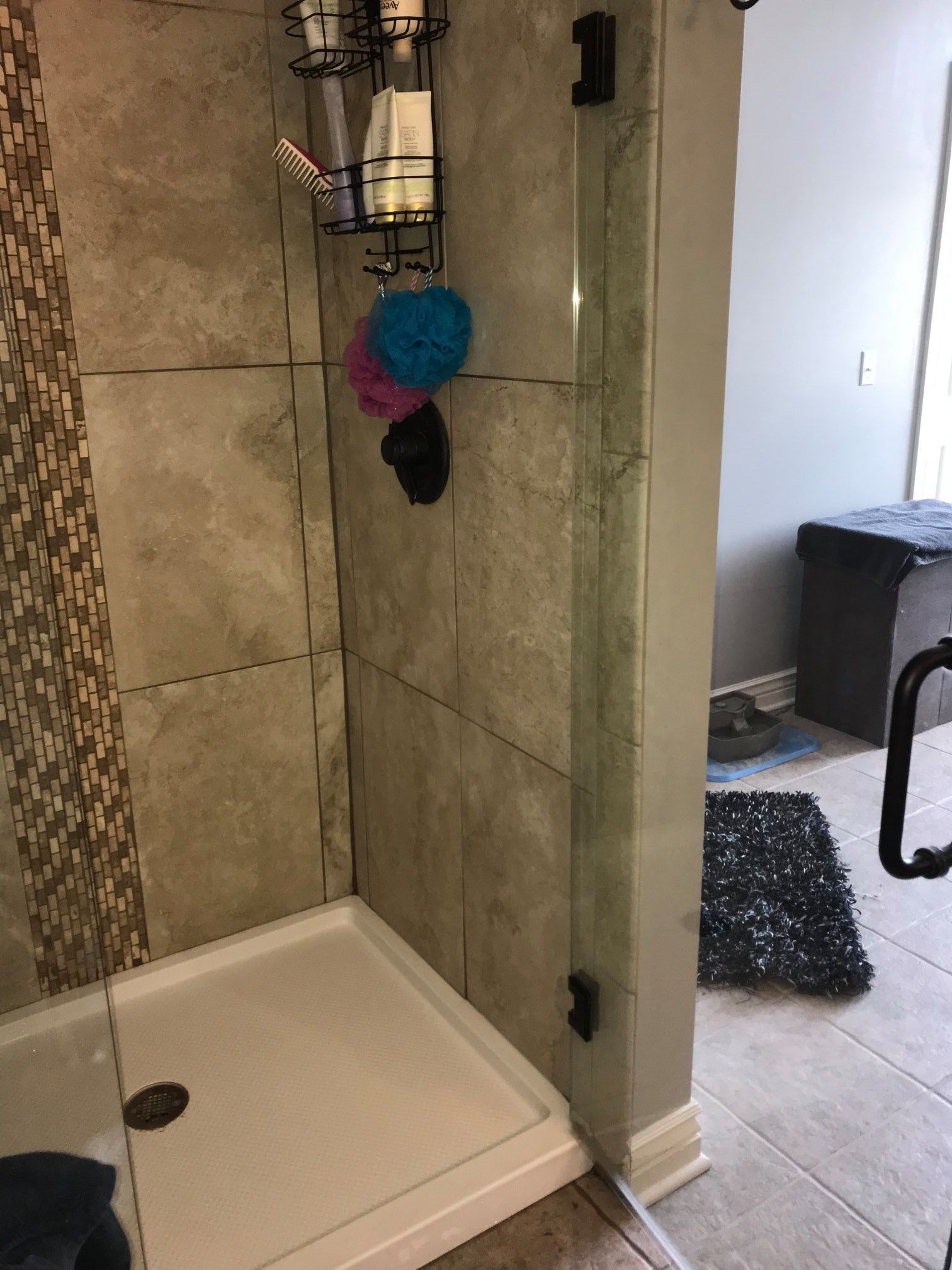 A shower with glass doors and gray tile walls. Soap and loofahs hang inside. A dark gray rug is on the floor outside.