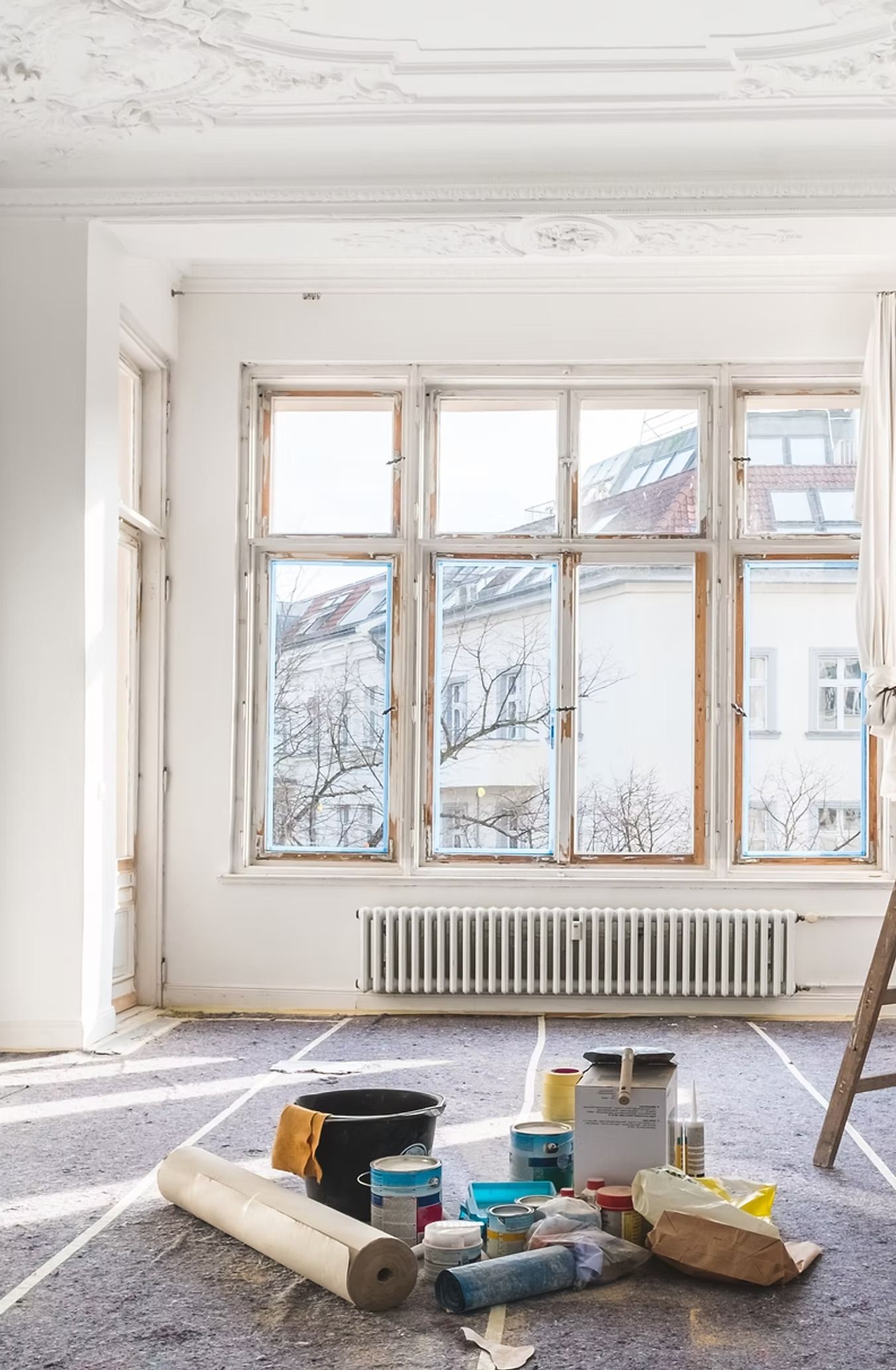 Renovation of a white room with window. Painting supplies on floor, ladder at side.