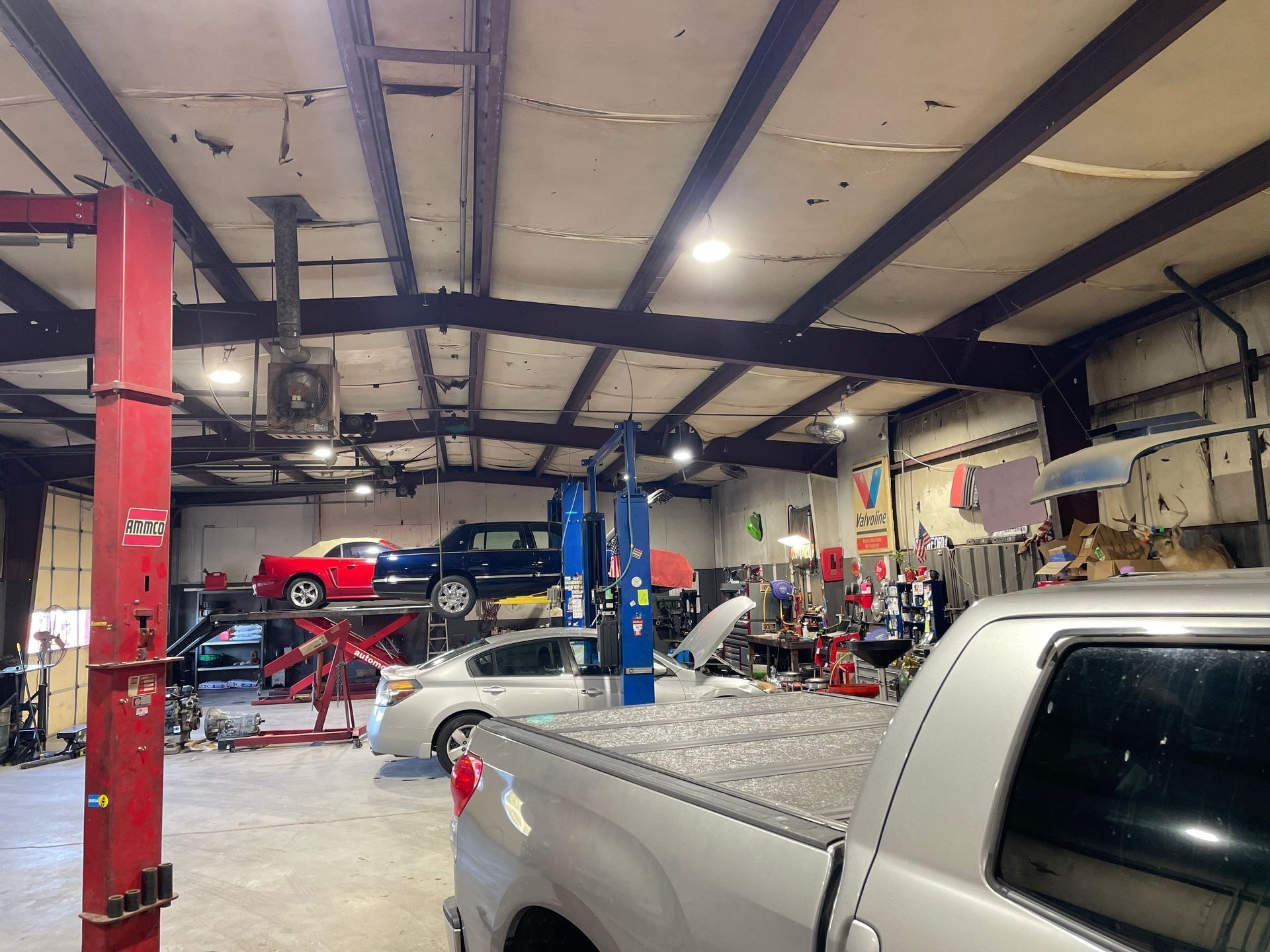 Interior of a busy auto repair shop. Cars on lifts, mechanics working, and a pickup truck parked inside.