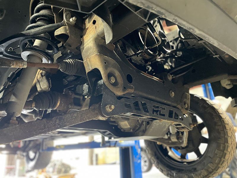 Underside view of a vehicle's front suspension. A black, modified suspension component is visible, along with a tire and other mechanical parts.