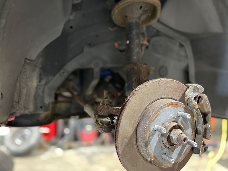 Car wheel assembly: brake rotor, caliper, suspension components, against car chassis. Rusty, worn appearance.