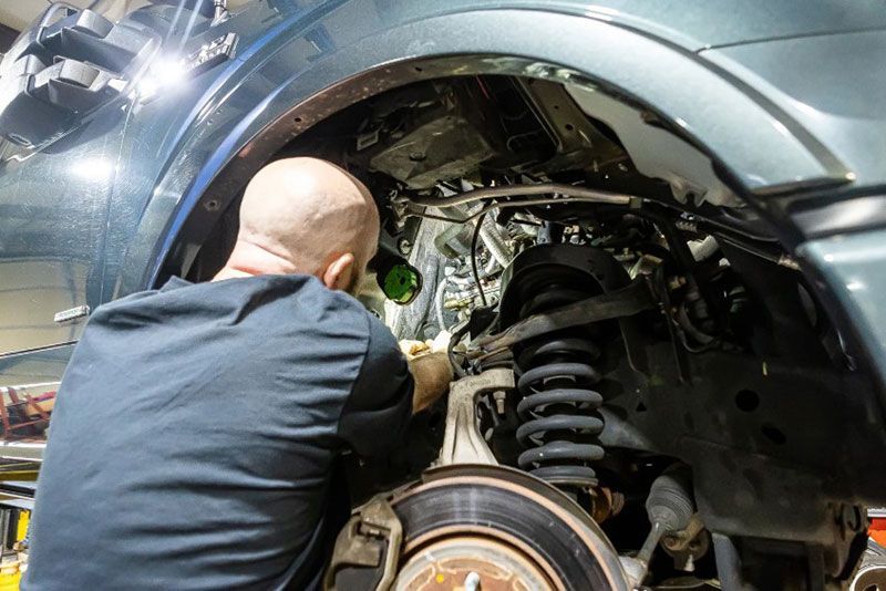 Mechanic working on the suspension of a blue vehicle in a garage, using a flashlight.