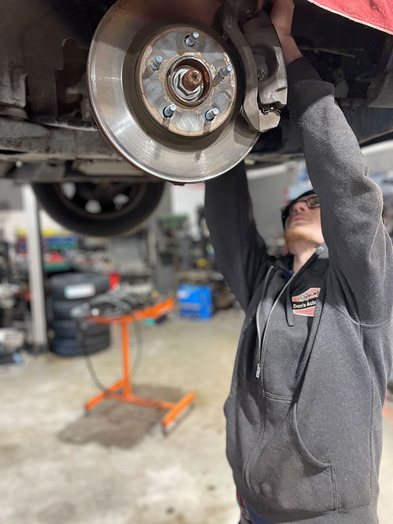 Mechanic working on car's brake system, viewed from underneath. Gray hoodie. Workshop setting.