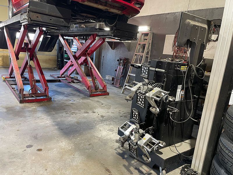 Car on a lift in a garage, alignment machine in foreground. Red lift, gray floor, tires and tools.