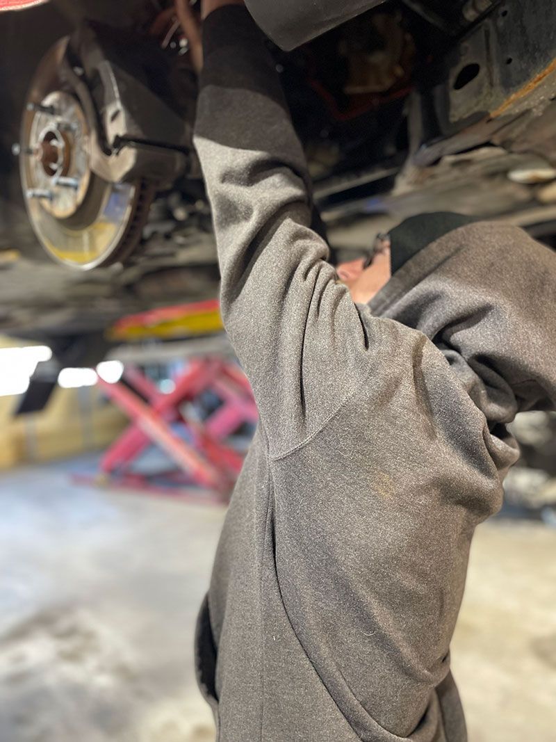 Mechanic working under a car, reaching up towards the undercarriage, wearing a gray hooded sweatshirt.