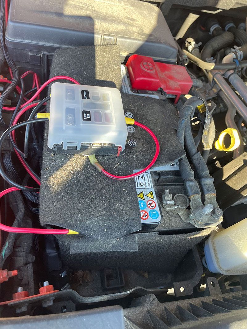 Car battery with a fuse box and wiring in an engine bay. Red and black wires.