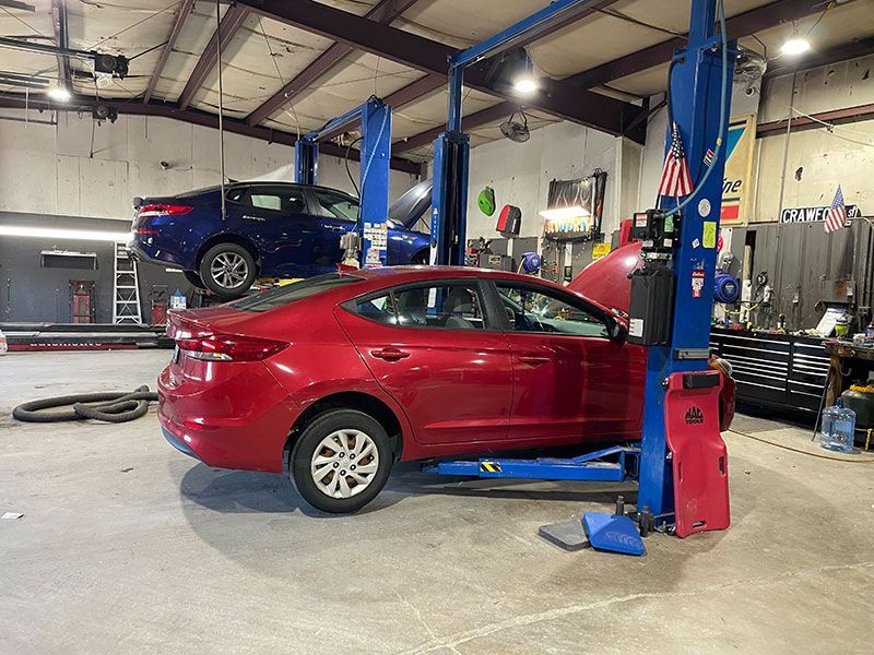 Red car on lift in auto repair shop; blue car above.