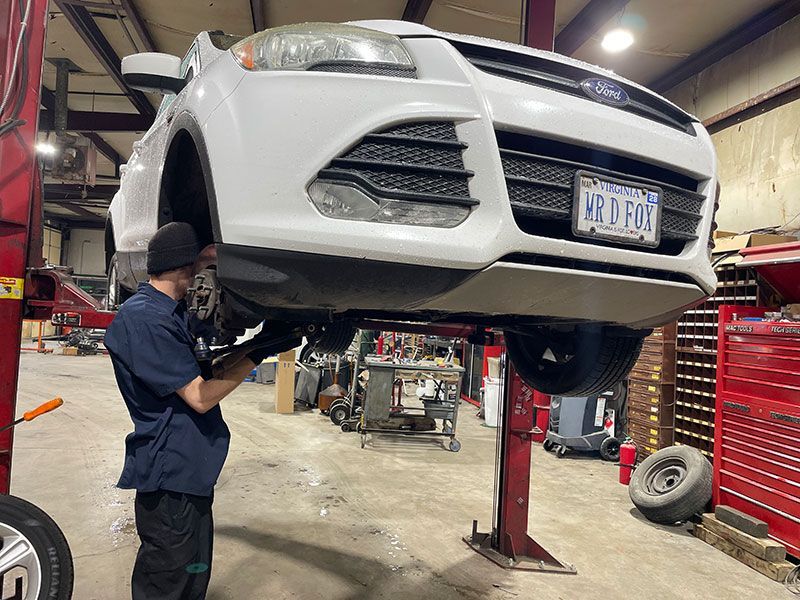 Mechanic working on a white Ford Escape raised on a lift in a garage.