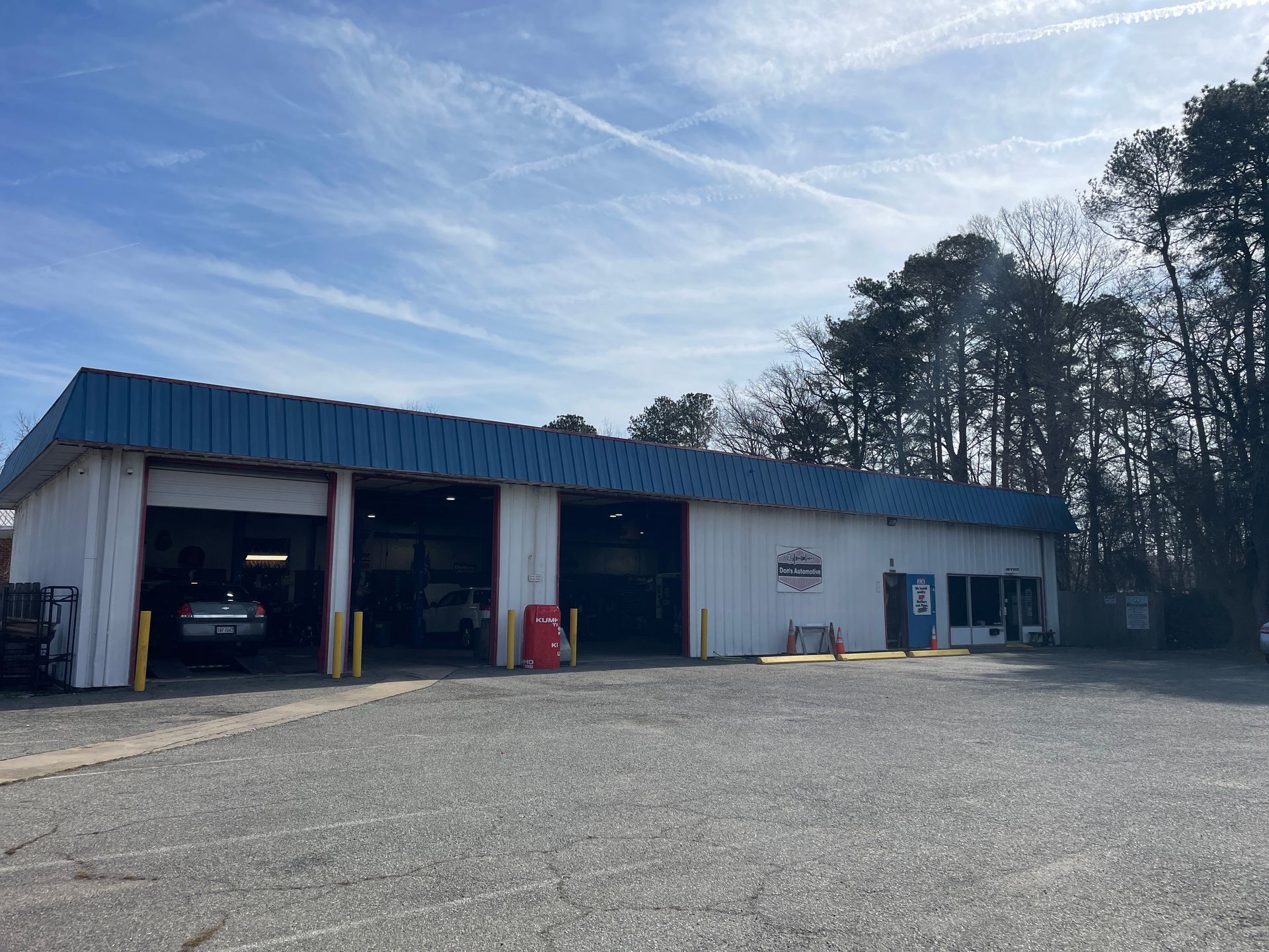 A white auto repair shop with three open garage bays and a blue metal roof, situated on a gravel lot under a sunny sky.