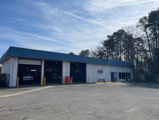 A white auto repair shop with a blue metal roof and three open garage bays stands in a gravel lot under a sunny sky.
