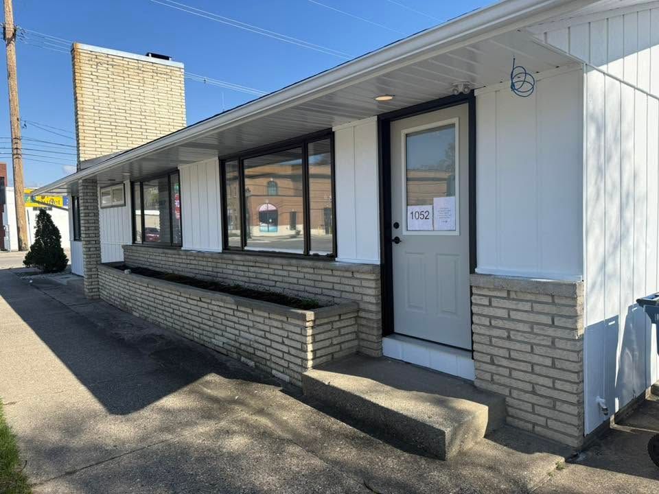 Small white storefront with a gray door, ramp, and large windows on a sunny street corner.