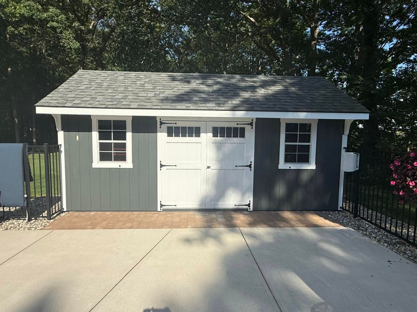Small gray shed with white double doors and windows beside a concrete driveway.