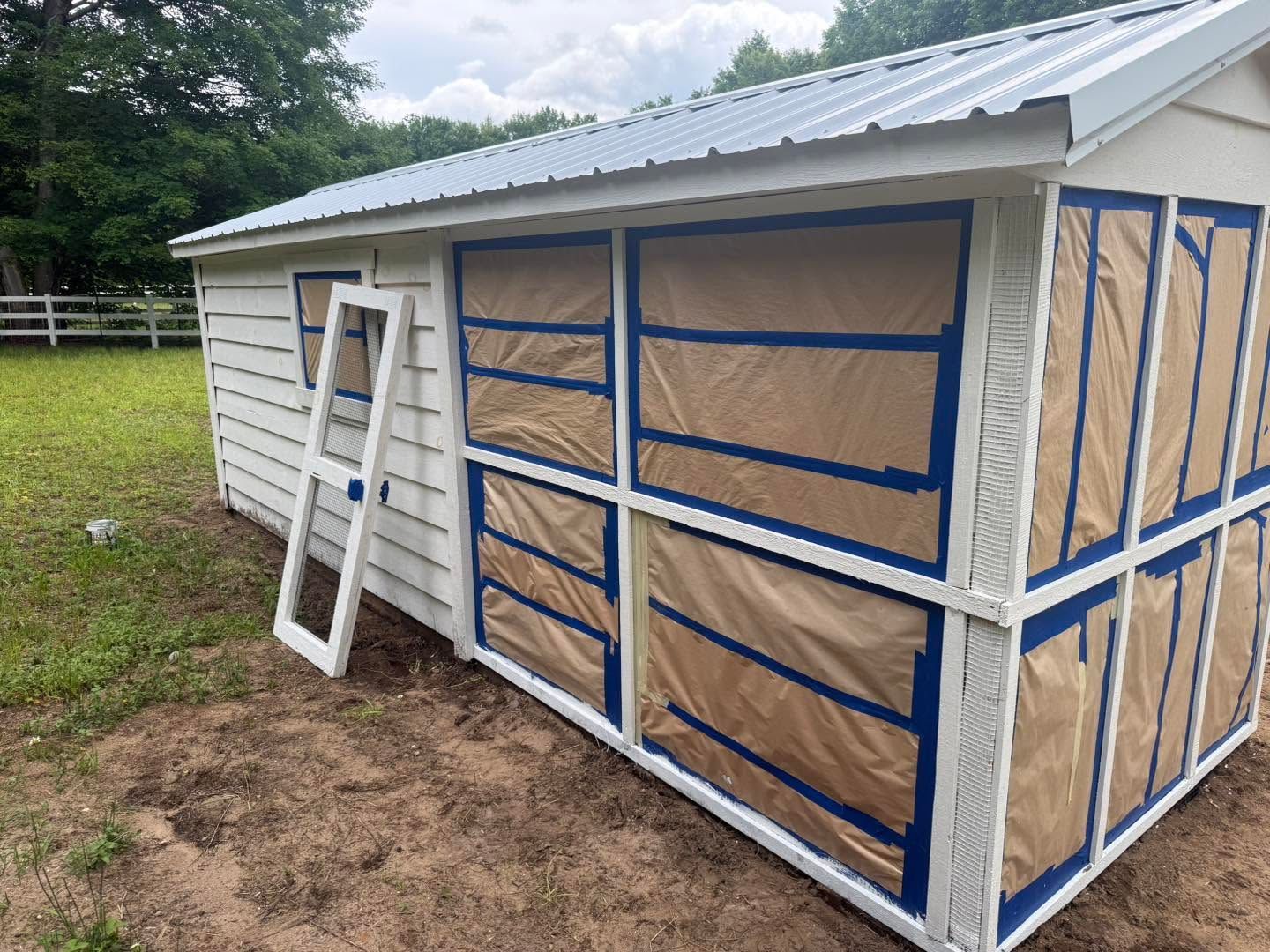 Partially wrapped white shed with blue tape and tan paper on the sides, outdoors on grass