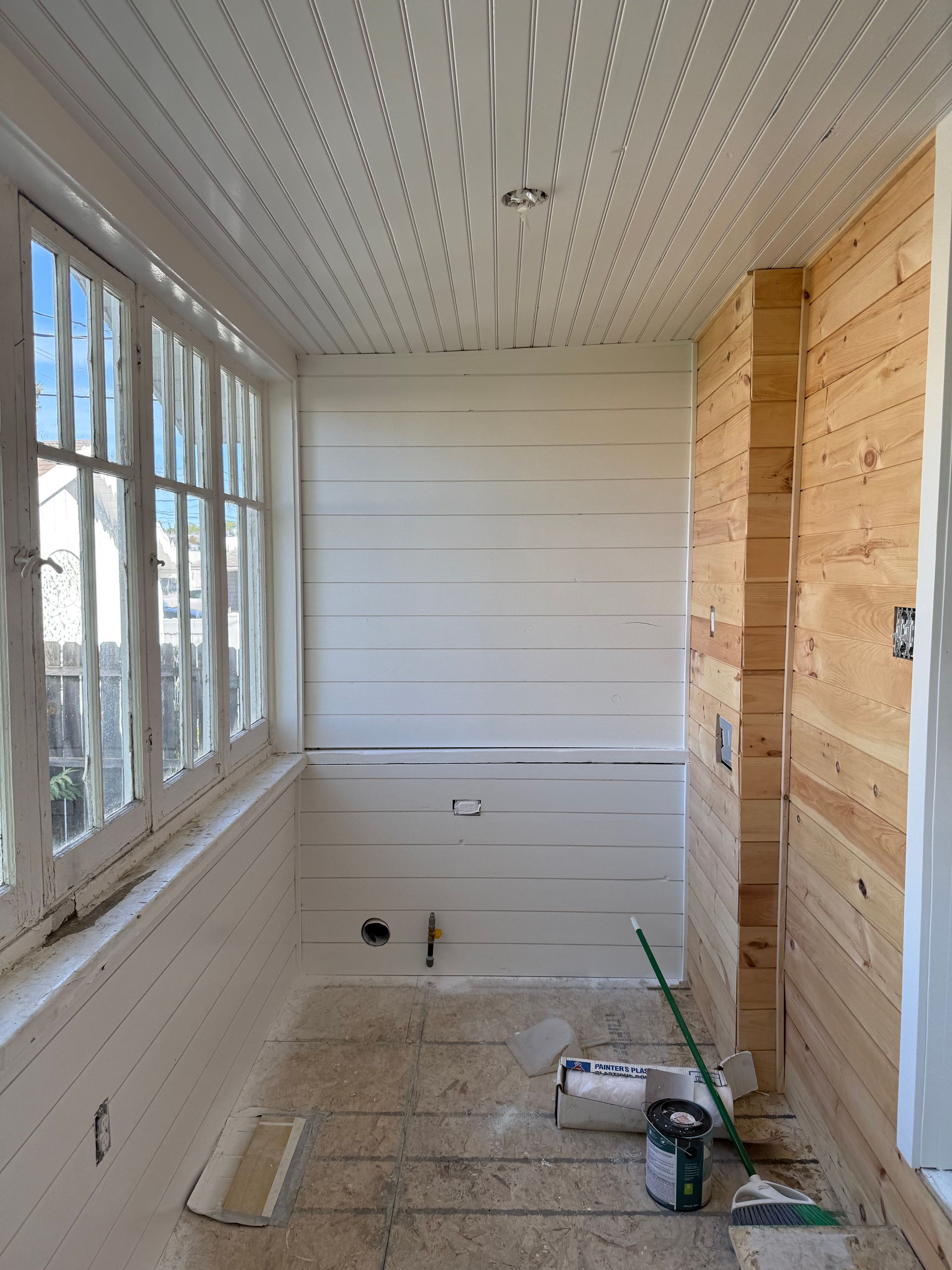 Narrow unfinished sunroom with white walls, wood paneling, large windows, and tools on the floor.