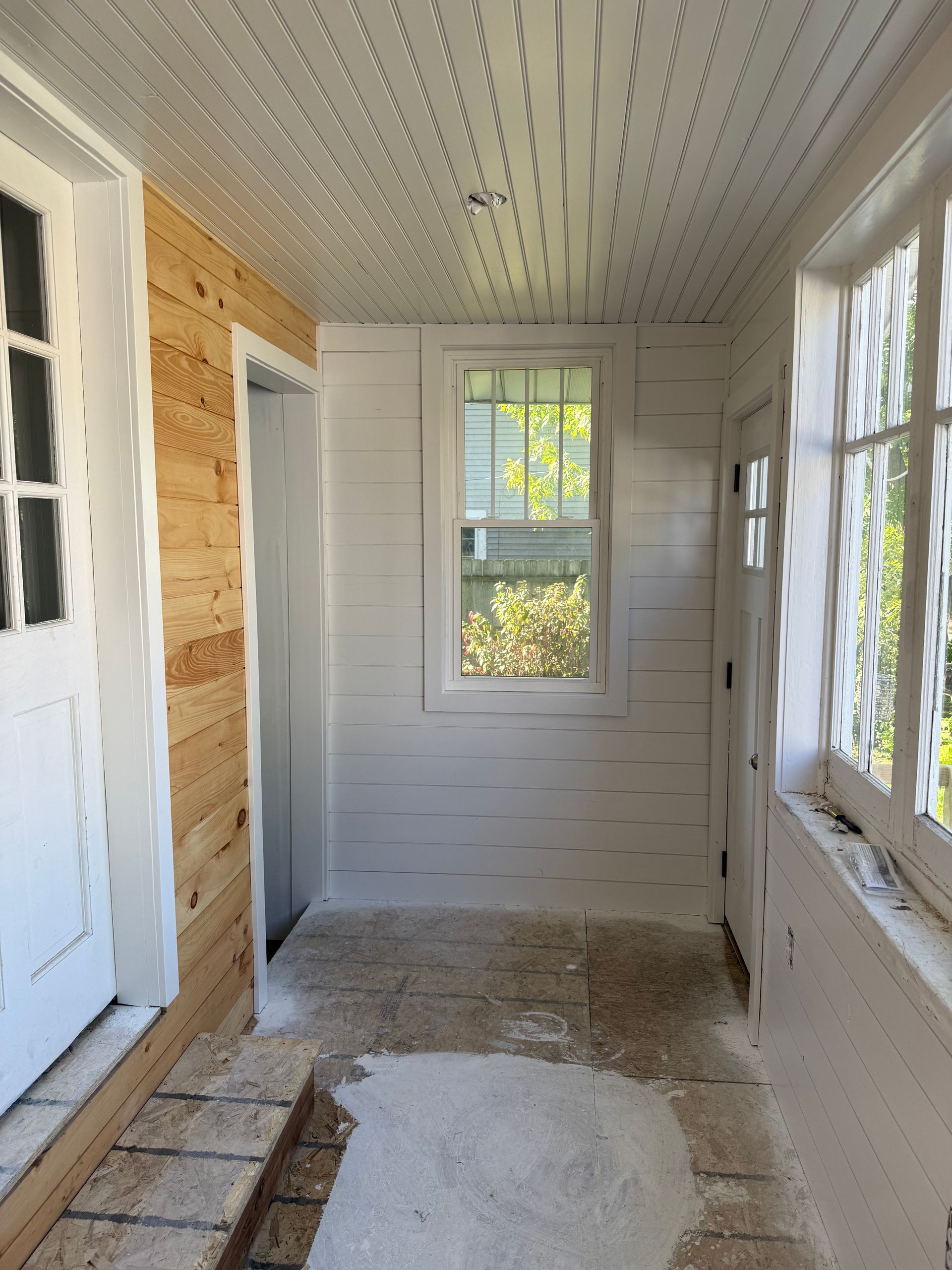 Narrow unfinished hallway with white walls, windows, and exposed floor patches under renovation