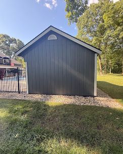 Gray shed with a gabled roof beside a black metal gate, surrounded by grass and trees