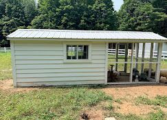 Small white shed with metal roof and screened porch in a grassy yard, backed by trees