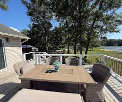 Deck with wicker dining table and chairs overlooking a lake beside a house on a sunny day