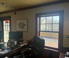 Home office with desk, computer, black chair, framed art, and a large window showing a deck and trees.