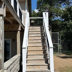 Outdoor wooden staircase with white railings beside a building, leading up to a fenced area.