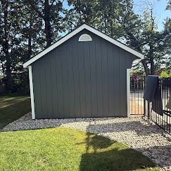Gray backyard shed with white trim, gravel base, and a black fence beside a grassy lawn