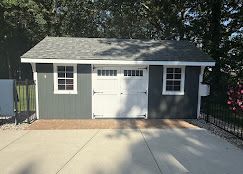 Small gray detached garage with white doors and windows, viewed from a driveway.