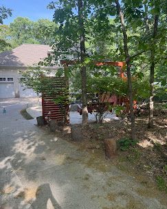 Wooded driveway beside a house with red patio furniture and a garage in the background.