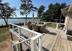 Wooden lakeside deck with patio chairs, railing, and trees overlooking the water