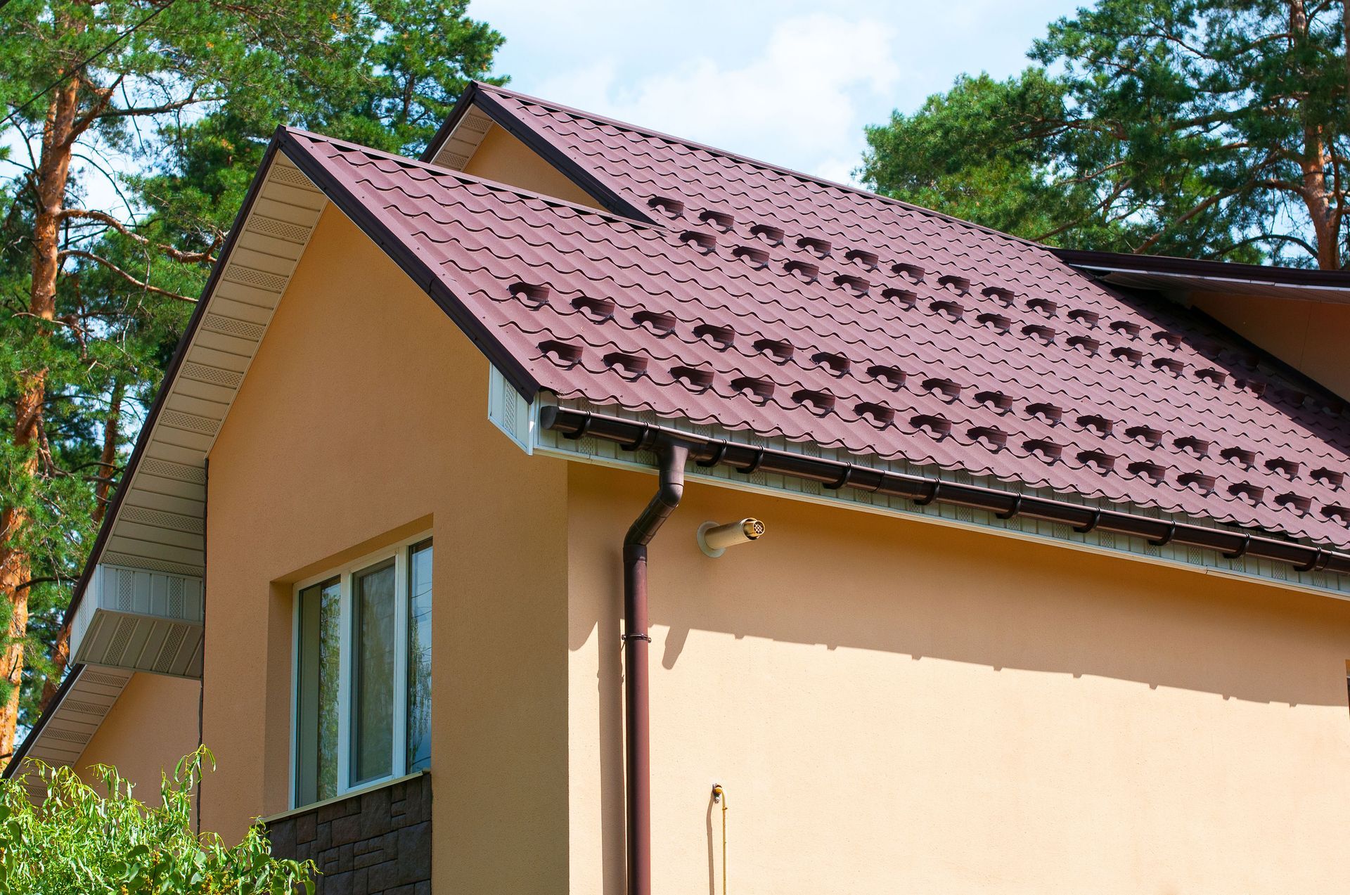 House with beige walls and a red-tiled roof, viewed from below against leafy trees and blue sky