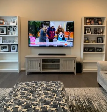 Living room with TV, white shelves of framed photos, and a patterned ottoman in the foreground