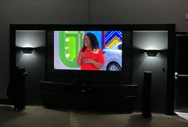 Large TV in a dark home theater displays a woman in a red shirt on a colorful set.