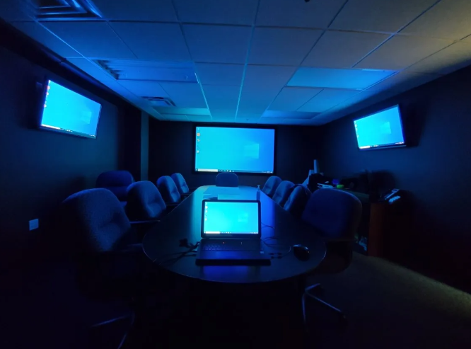 Dark conference room with blue-lit screens and a laptop on a table, viewed from the back of the room