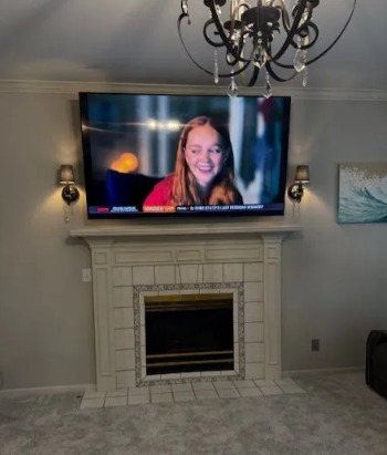 TV mounted above a fireplace shows a smiling woman during a video call in a living room.