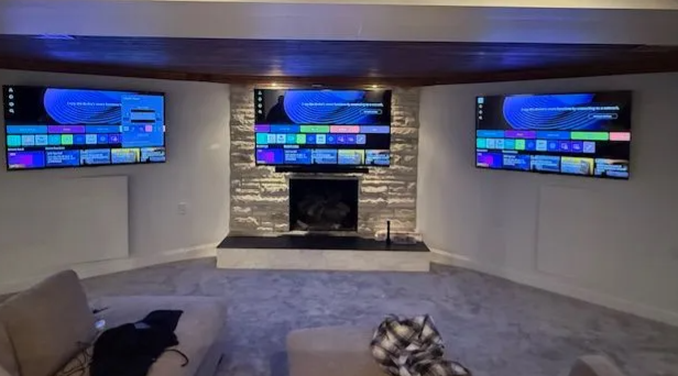 Living room with three wall-mounted TVs above a stone fireplace and a gray carpeted seating area