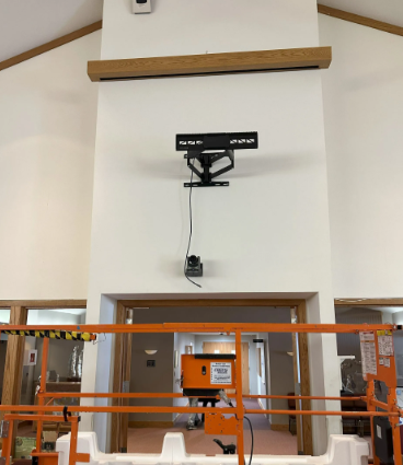 Wall-mounted projector above a doorway in a bright indoor room, with orange scaffolding in front