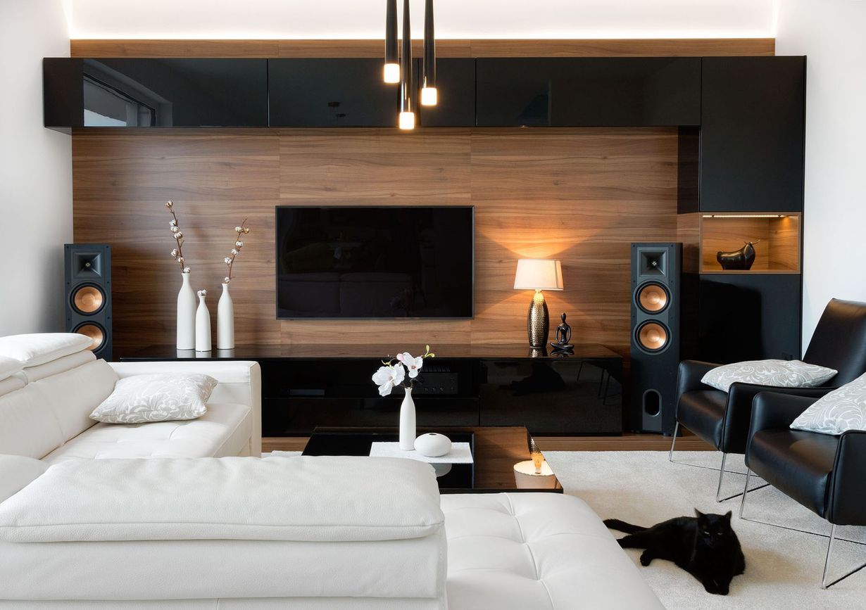 Modern living room with black sectional, white ottoman, wall TV, wood paneling, and speakers