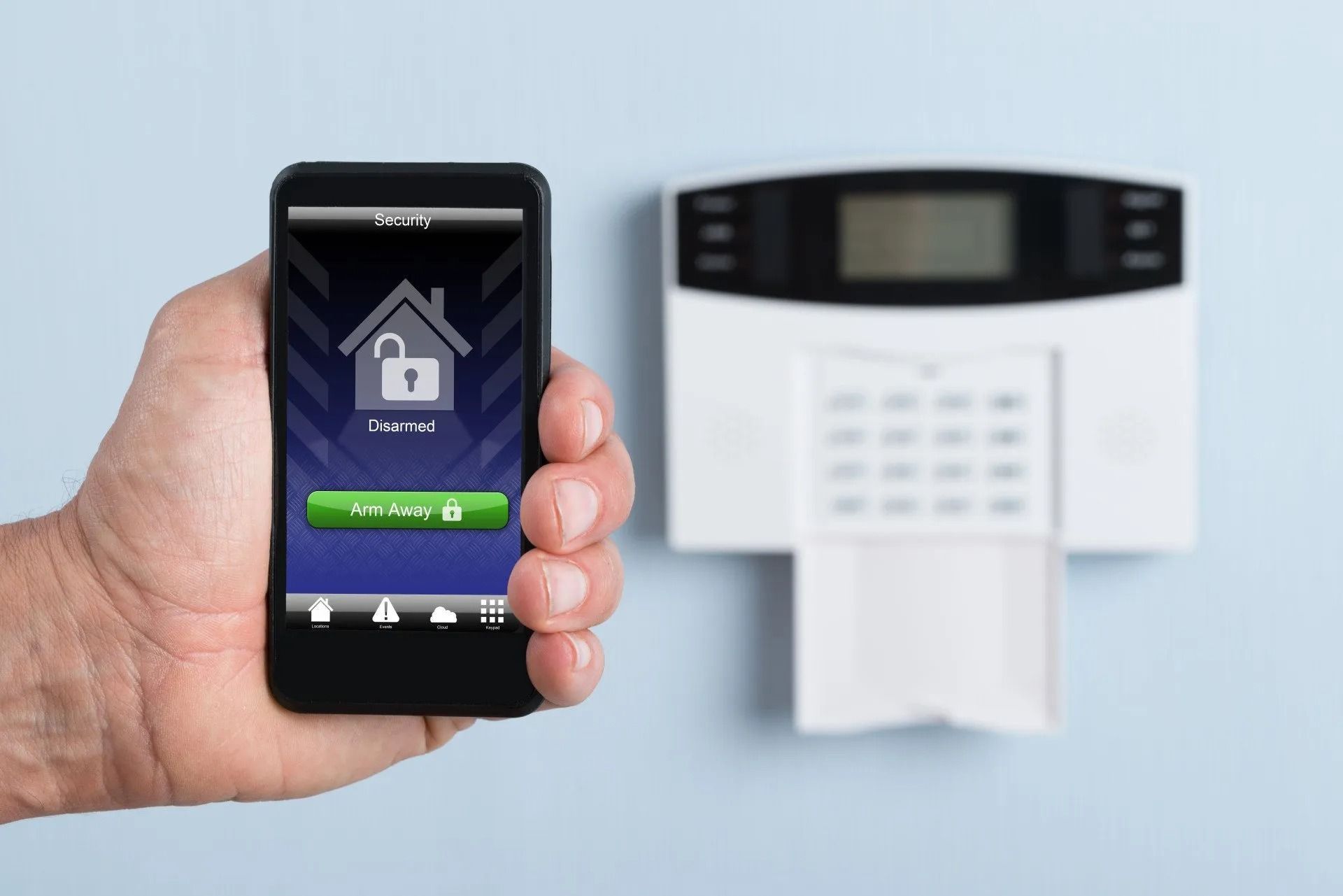Hand holding smartphone with home app next to a wall-mounted security system keypad