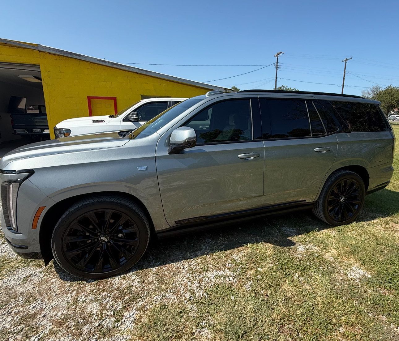 Gray Cadillac Escalade SUV with black wheels parked on grass in front of a building under a blue sky.