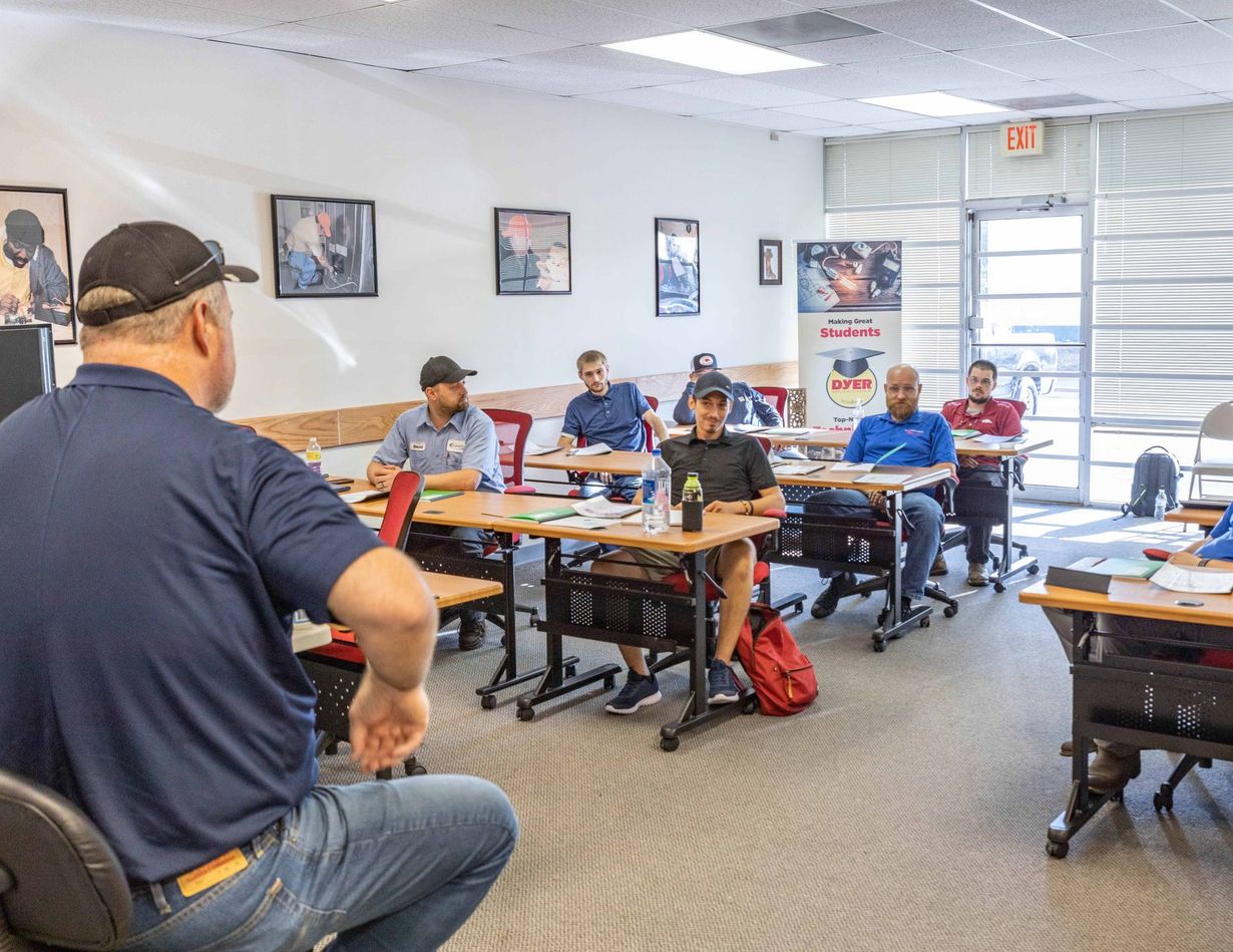 Instructor in a classroom teaching seated students. White room with desks, posters, and an exit door.