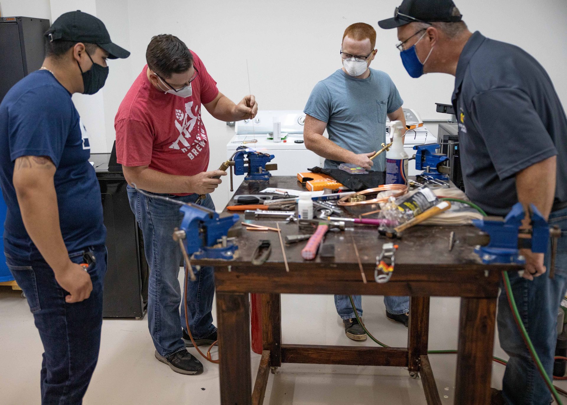 Four people wearing masks work at a workbench with tools. They appear to be repairing something.