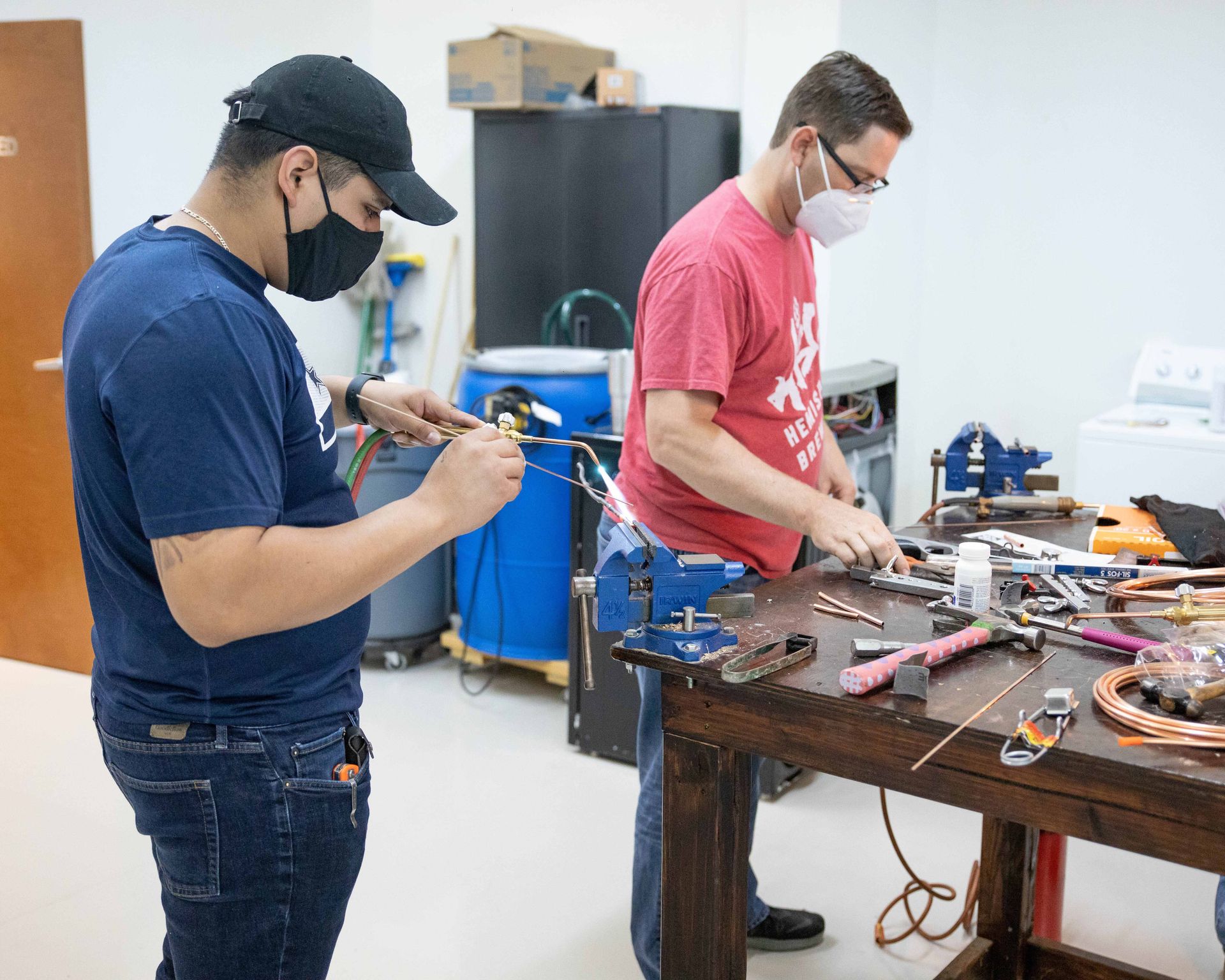 Two people working at a workbench, one wiring, the other assembling parts, both wearing masks.