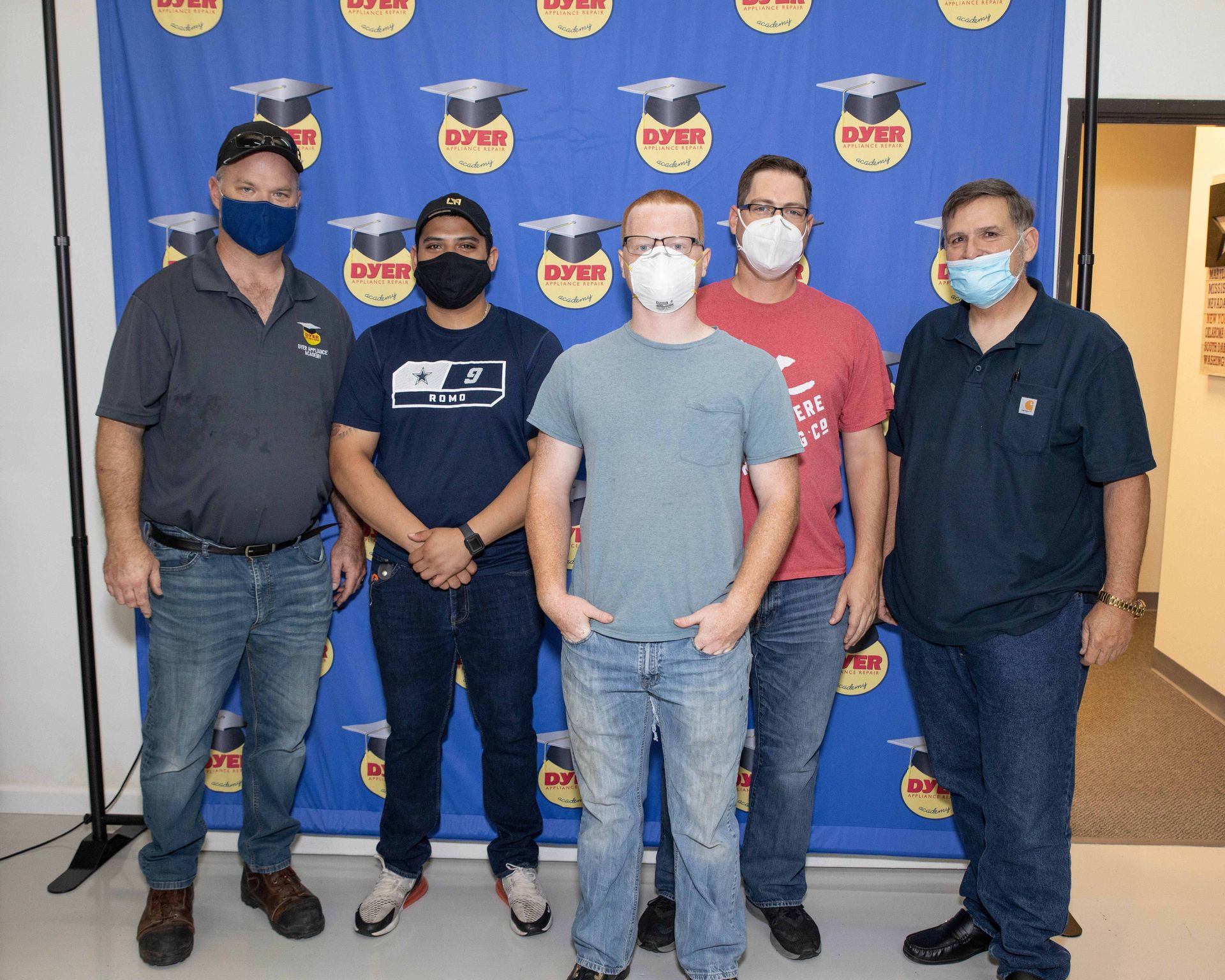Five people wearing masks pose in front of a blue backdrop with graduation caps.