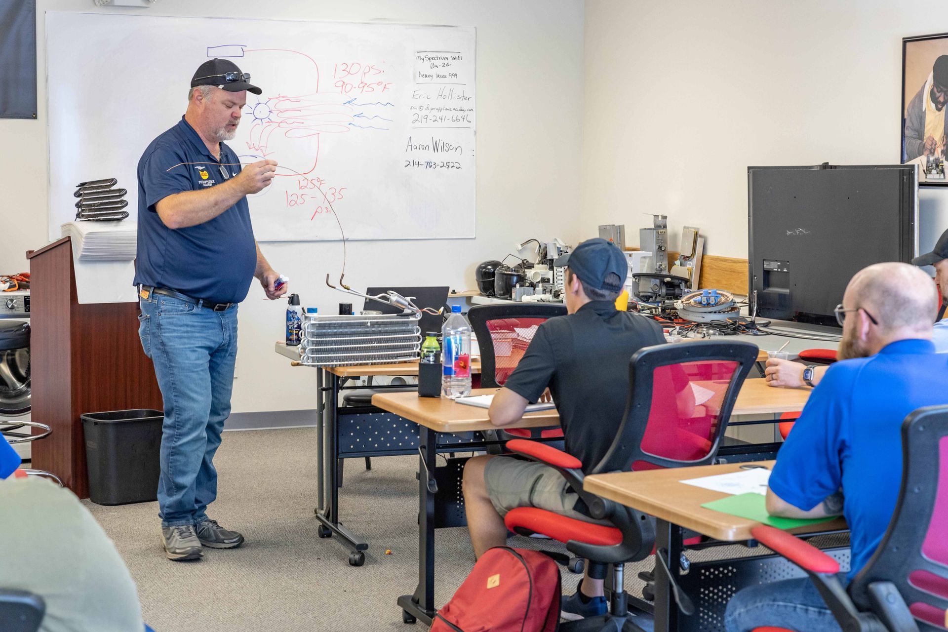 Man lecturing in a classroom with a whiteboard, students at desks.