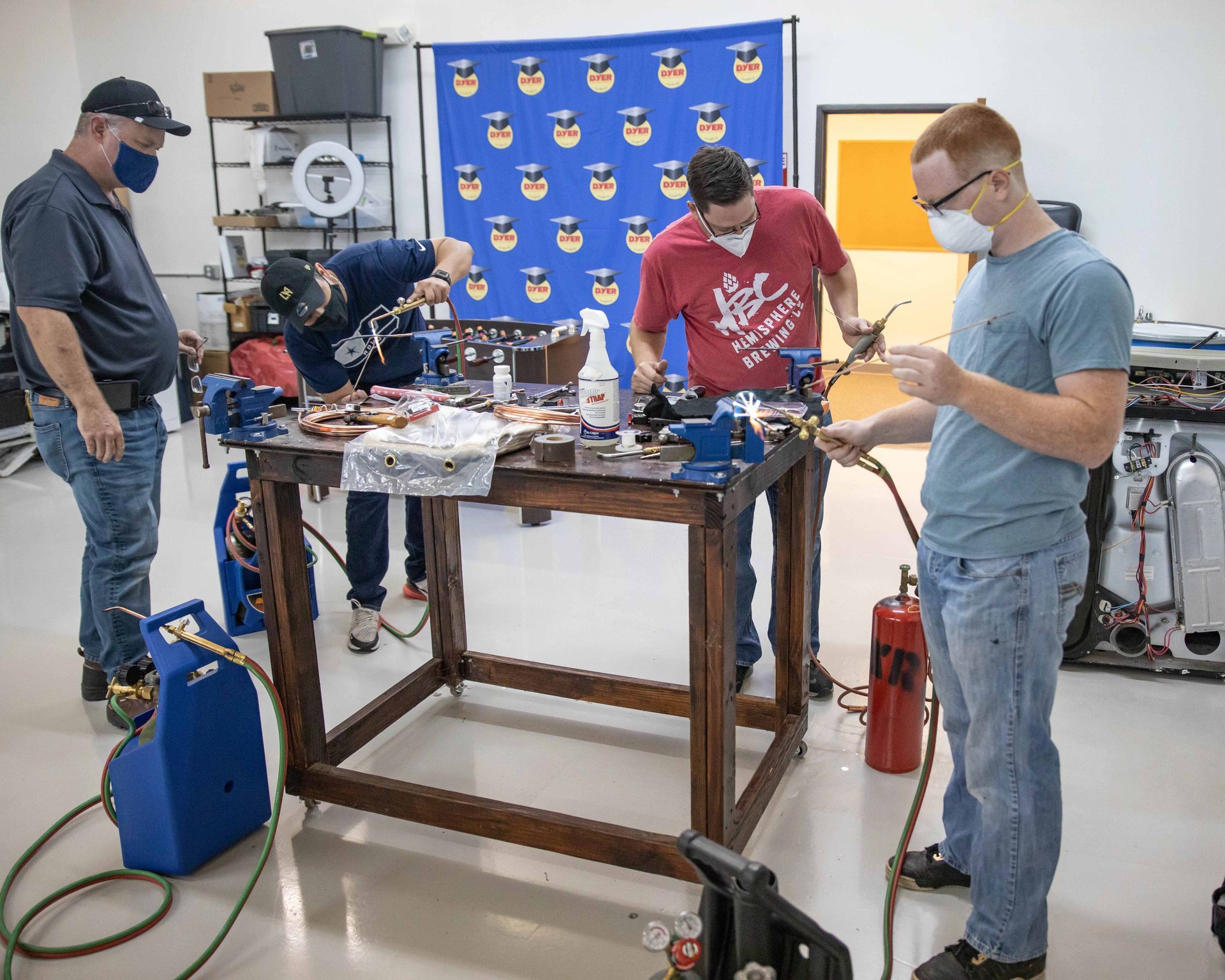 Four people wearing masks work on a project at a workbench. Tools and wires cover the surface.