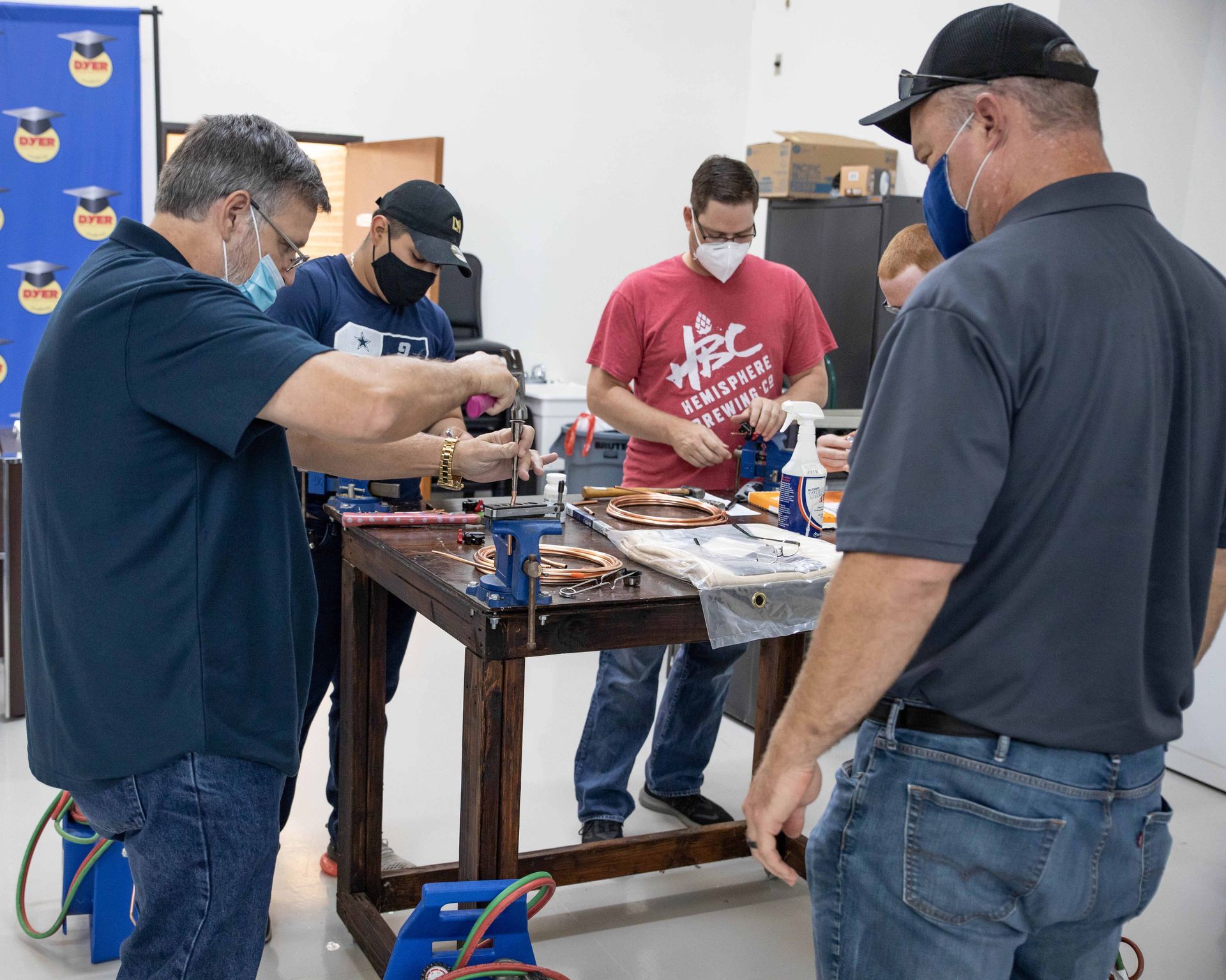 People working on a table with tools. Indoors, four men wearing masks, a child. Copper piping and wires visible.