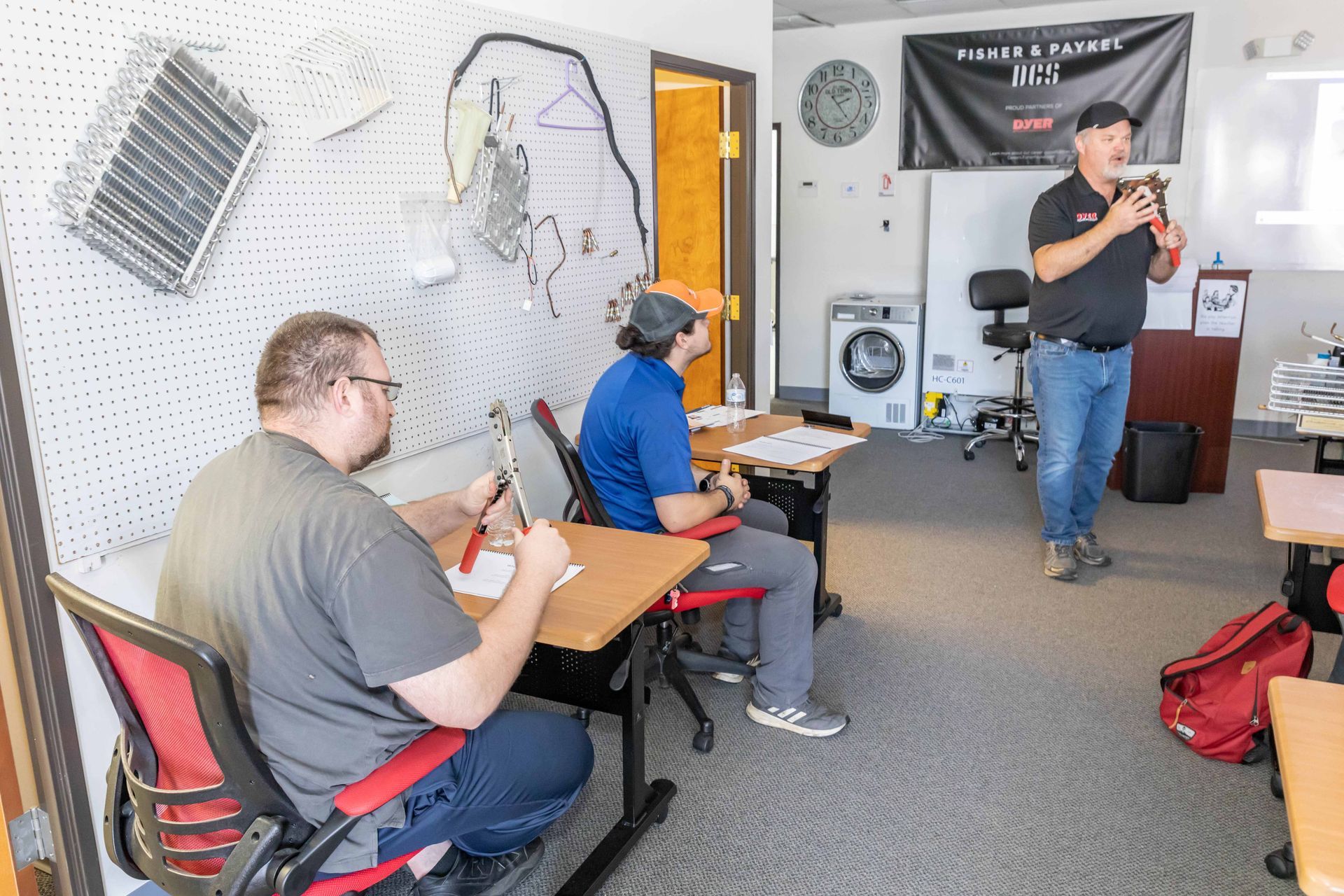 A man teaches a class; two students take notes. Classroom setting with desks and a pegboard.