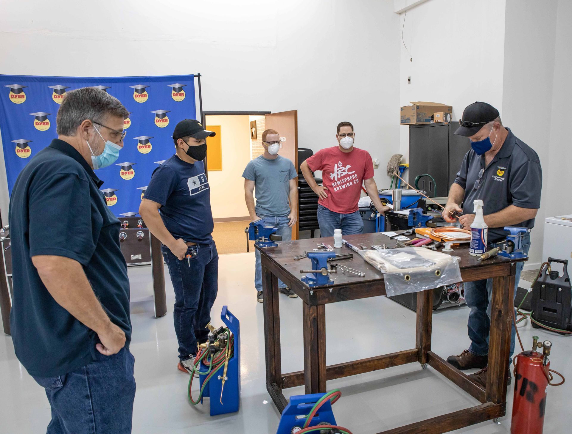 Men in masks observe a workshop demonstration at a table with tools. A blue banner is in the background.