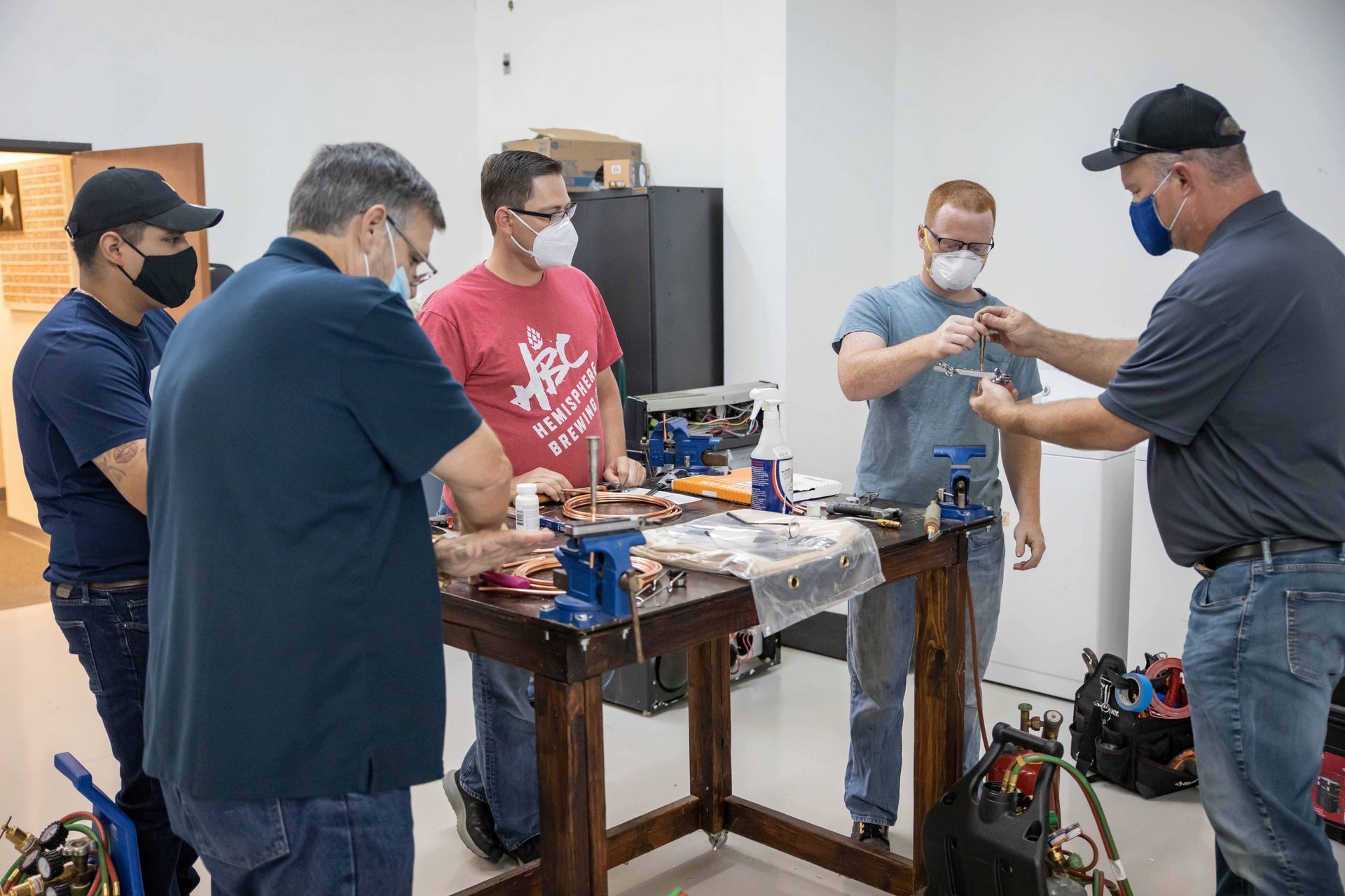 Group of masked people at a workshop, examining objects at a table. Workshop interior, tools visible.